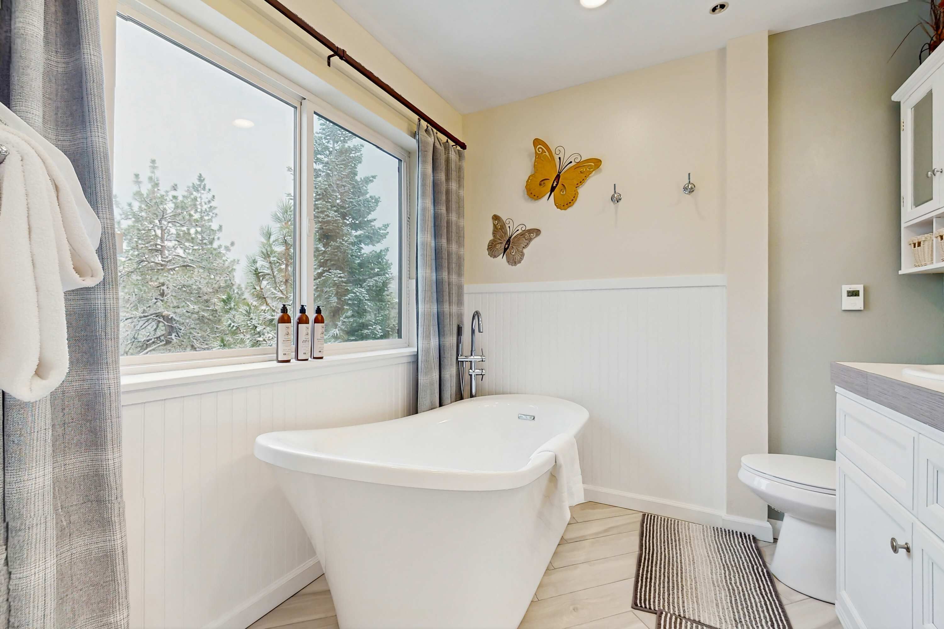 A bathroom with a freestanding bathtub, a toilet, a window with curtains, and wall-mounted butterfly decorations.