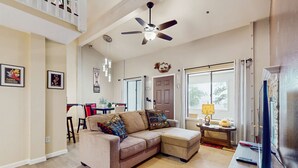 A living room with a sofa, ottoman, ceiling fan, television, and a dining area in the background.