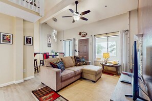 A living room with a sofa, ottoman, ceiling fan, television, and a dining area in the background. - Updated Condo with Lake Views, Heated Floors, Soaking Tub (Stateline)