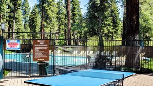 A ping pong table on a deck with a pool in the background, surrounded by a fence and trees. - Charming Condo with Shared Hot Tub, Pool, Resort Amenities - Close to Ski & Beach (Carnelian Bay)