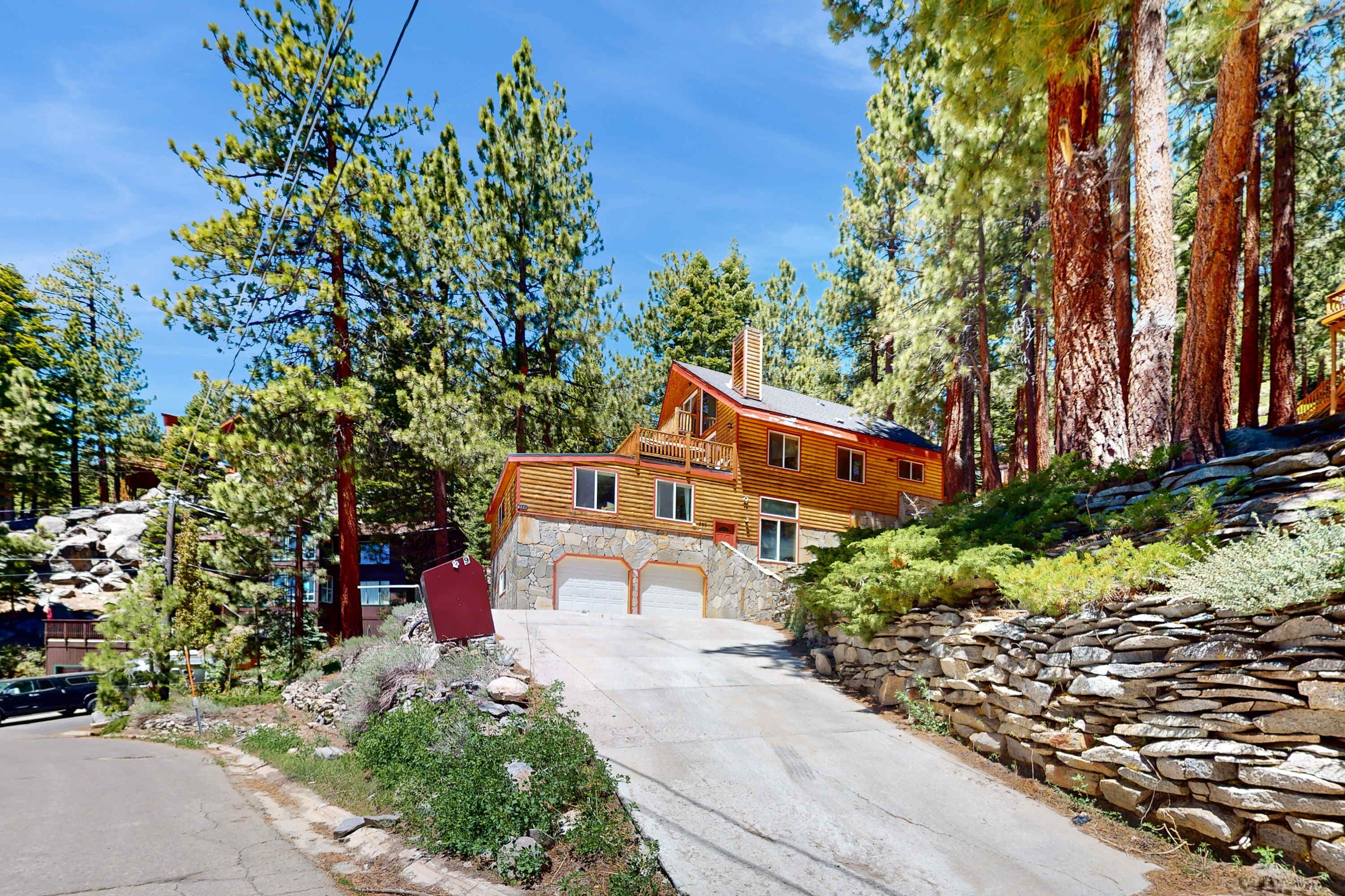 A two-story log cabin with a stone foundation, a stone retaining wall, and a driveway leading to a garage.