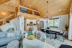 Un vaste espace de vie comprenant un canapé, un pouf et une table à manger, une cuisine en arrière-plan et un escalier en bois.