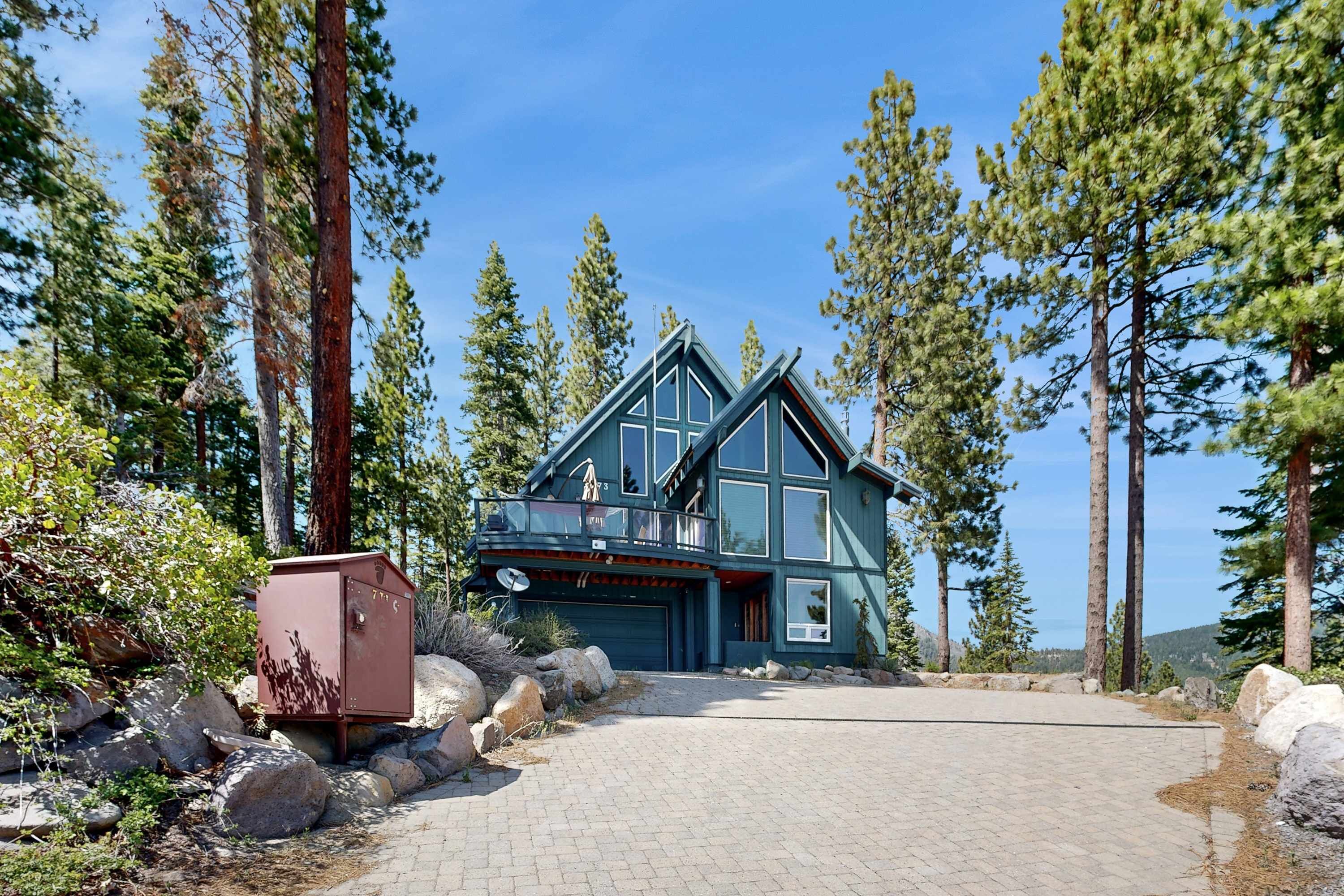 A two-story house with a large garage, surrounded by tall pine trees and a paved driveway.