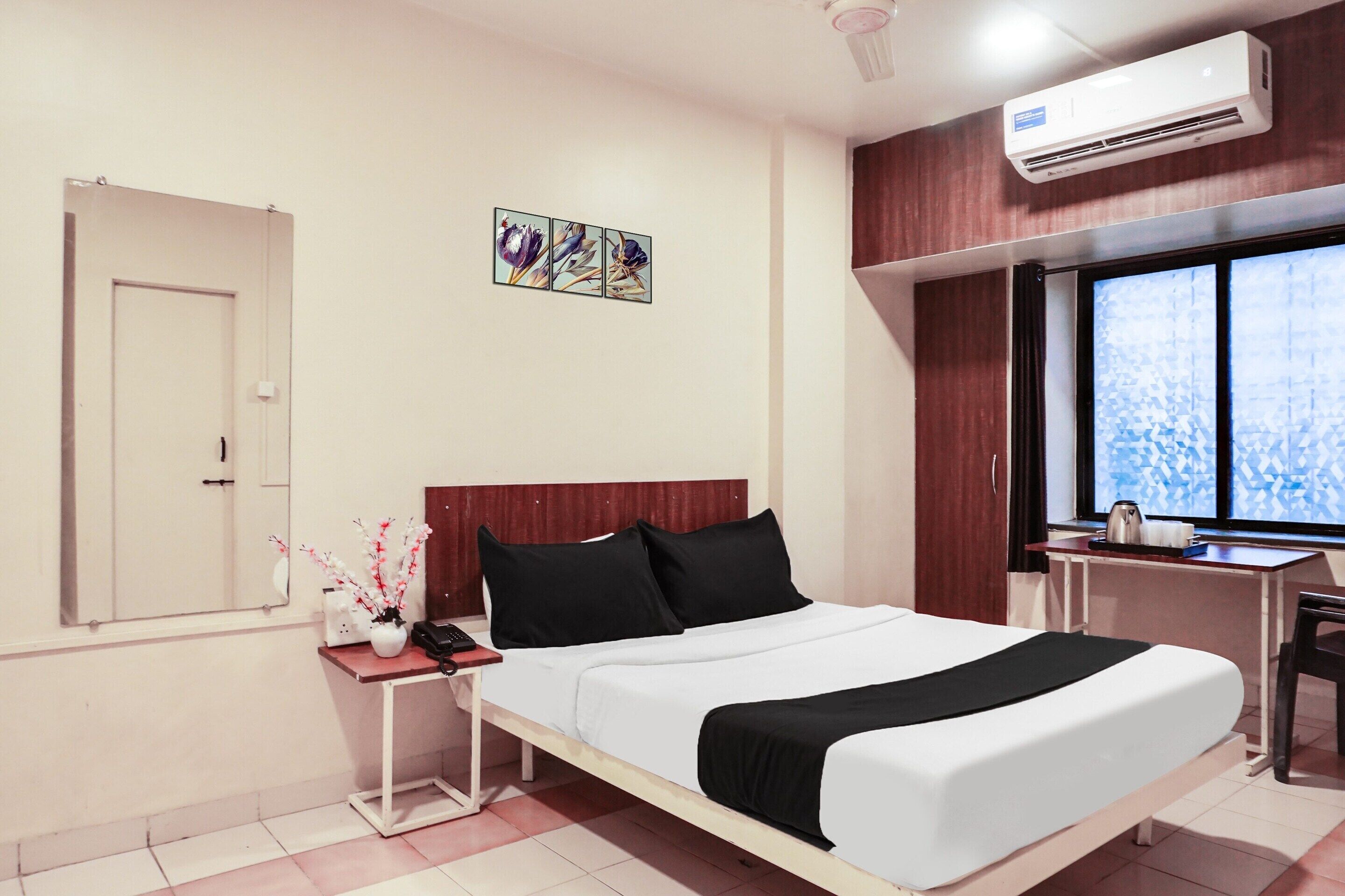 A hotel room with a bed, bedside table, air conditioning unit, window, and artwork on the wall.