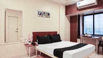 A hotel room with a bed, bedside table, air conditioning unit, window, and artwork on the wall.