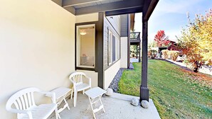 A covered outdoor seating area with two white plastic chairs and a small table, a concrete floor, and a view of a residential street with houses and trees. - Quiet Townhouse in Ideal Truckee Location with Pool, Hot Tub & Sauna (Truckee)
