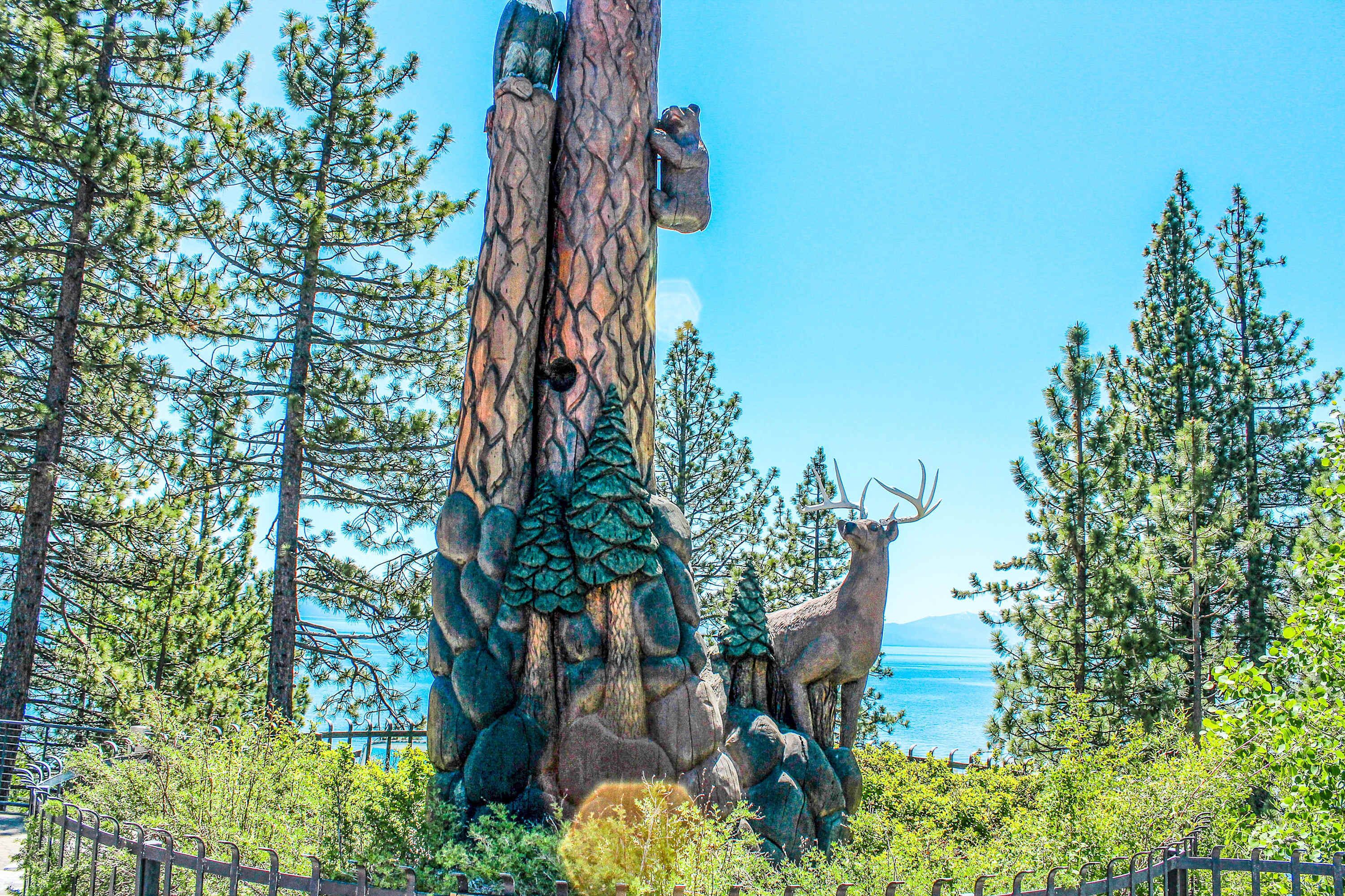 Eine große Baumskulptur mit einem Bären und einem Reh, umgeben von Kiefern und blauem Himmel.