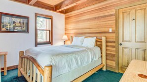 Ein Bett mit Holzkopfstütze, ein Nachttisch, ein Fenster und eine Tür in einem Zimmer mit Holzwänden und -decke.