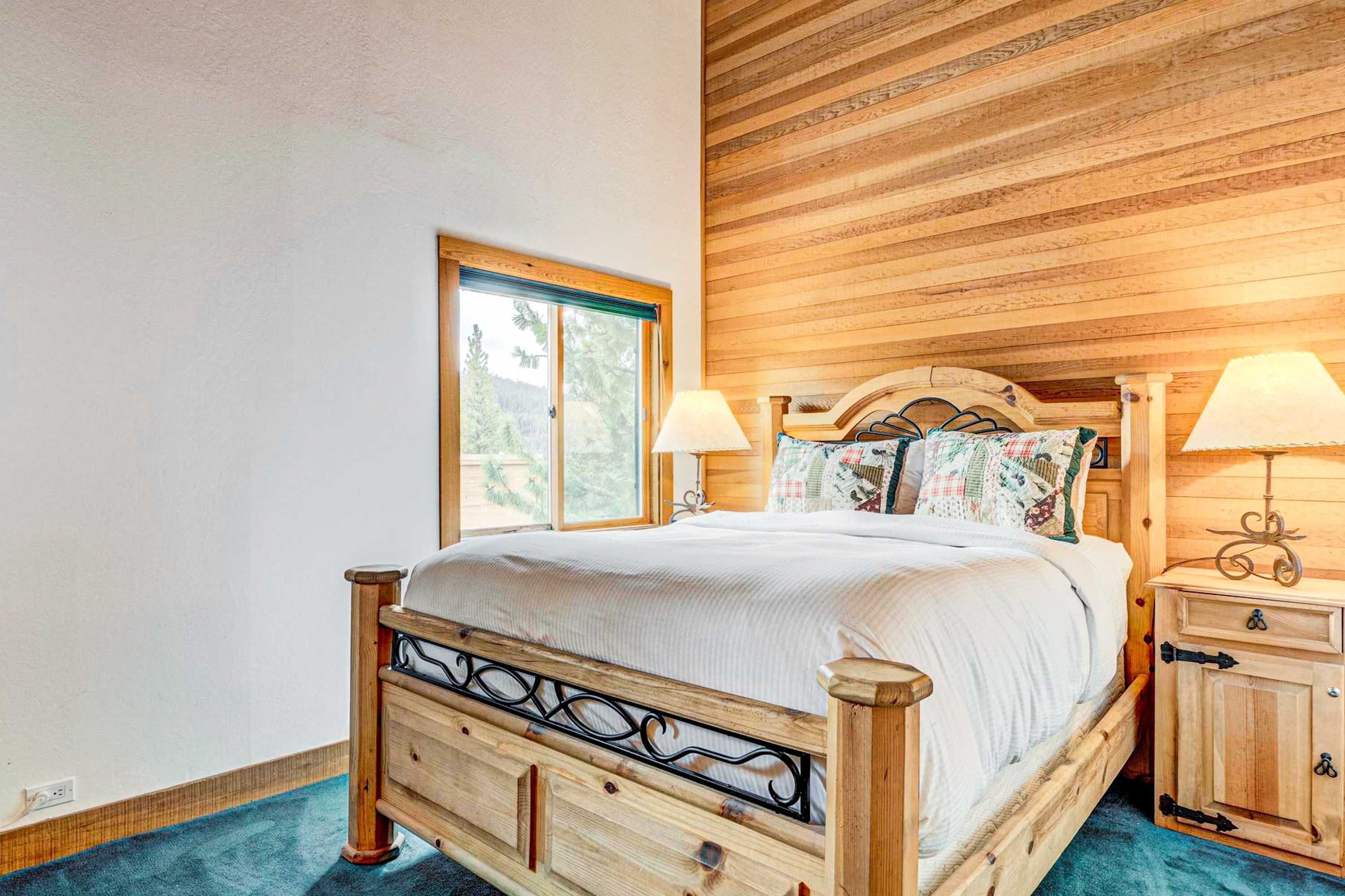 Una cama de madera con cabecera, dos lámparas de noche, una mesita de noche y una ventana con vista a árboles.