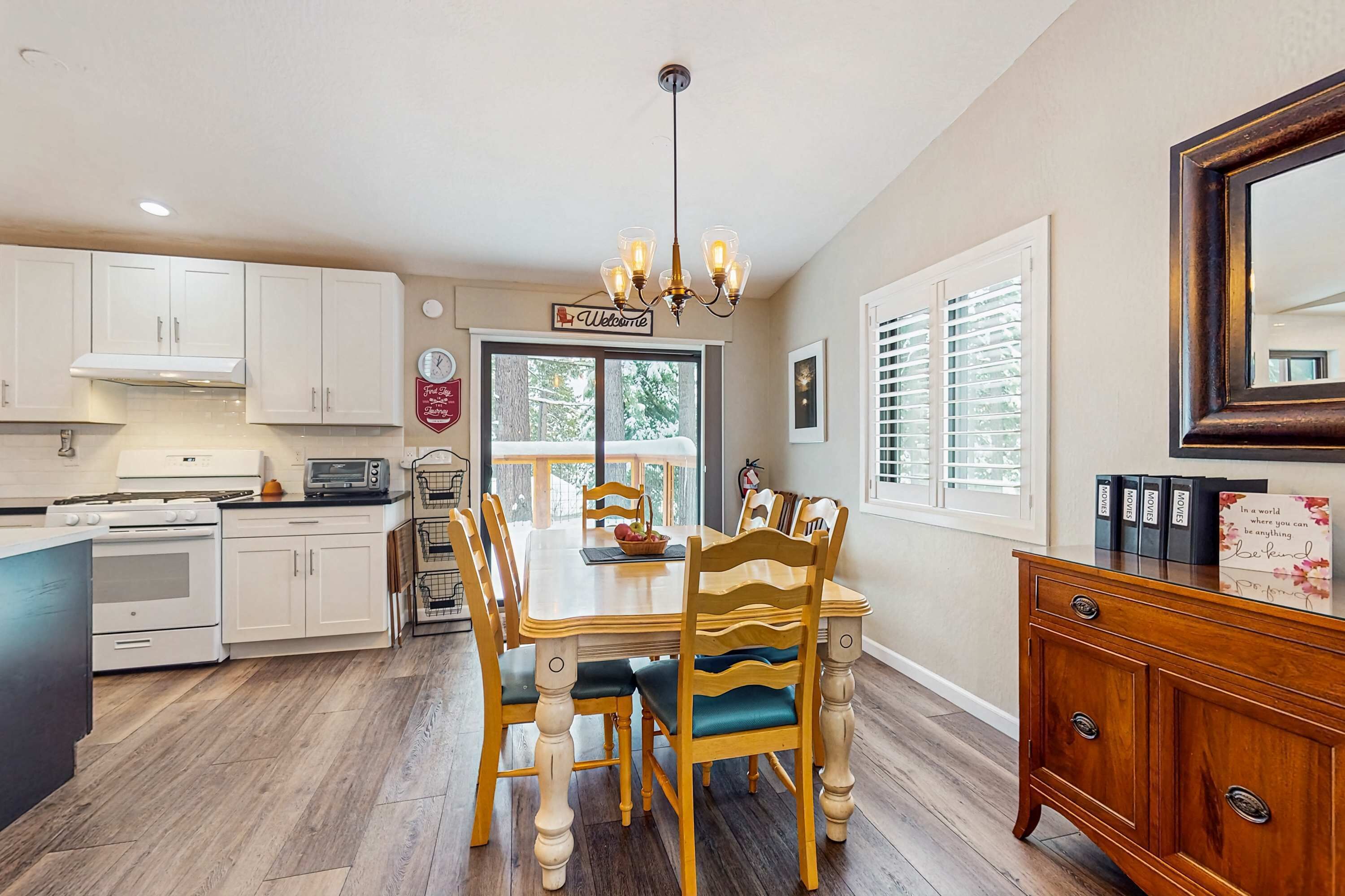 A kitchen with white cabinets, a dining table with wooden chairs, a chandelier, and a view through the window.
