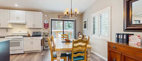 A kitchen with white cabinets, a dining table with wooden chairs, a chandelier, and a view through the window.