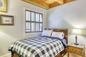 Ein Schlafzimmer mit einem Bett, einem Nachttisch, einer Lampe, einem Fenster mit Jalousien und zwei gerahmten Bildern an der Wand.