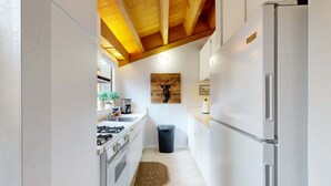 A narrow kitchen with white cabinets, a stainless steel refrigerator, a wooden ceiling, and a trash bin. - Mountain Condo with Fireplace, Balcony, WiFi (NorthStar-Truckee)