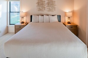 Een groot bed met een witte dekbedovertrek, twee nachtkastjes met lampen, een kunstwerk aan de muur en een raam met uitzicht.