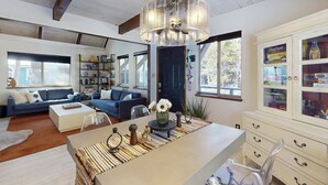 A spacious living room with a large window, a blue sofa, a coffee table, and a dining area with a clear glass chair. - Family-friendly cabin with fireplace, bikes, loft, games, & skiing nearby (South Lake Tahoe)