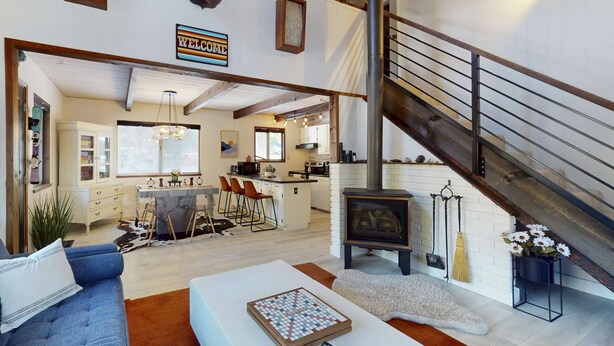 A spacious living area with a sofa, a coffee table, a fireplace, a kitchen in the background, and a staircase. - Family-friendly cabin with fireplace, bikes, loft, games, & skiing nearby (South Lake Tahoe)