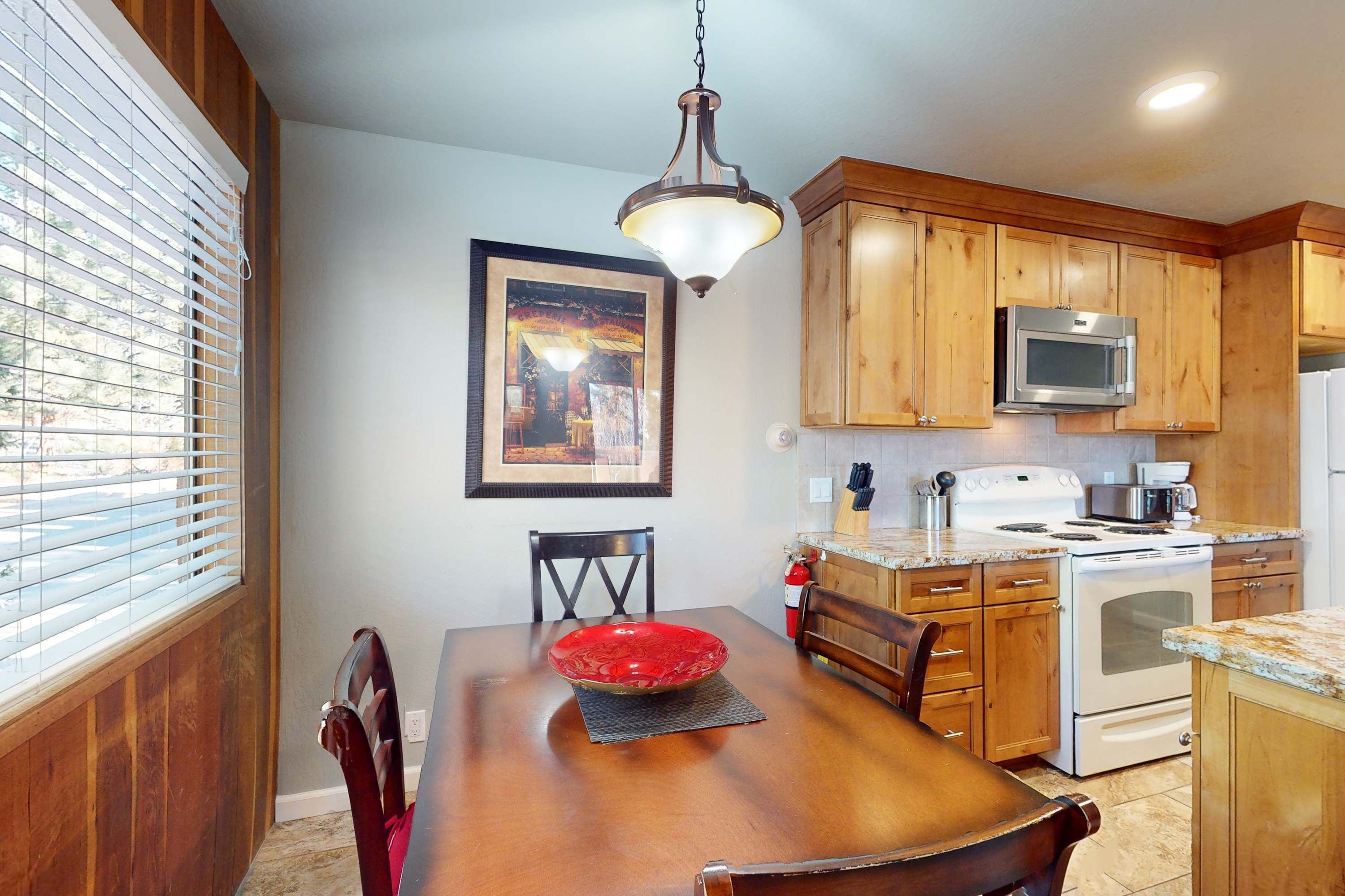 Une table à manger avec des chaises, un bol rouge, un tableau encadré, des meubles en bois, un four à micro-ondes et une cuisinière.