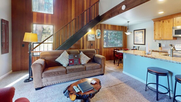 A spacious living area with a wooden staircase, a sofa with cushions, a coffee table, and a kitchen area with a bar. - Beautiful Condo with Shared Pool & Tennis Courts, Close to Northstar (Kings Beach)