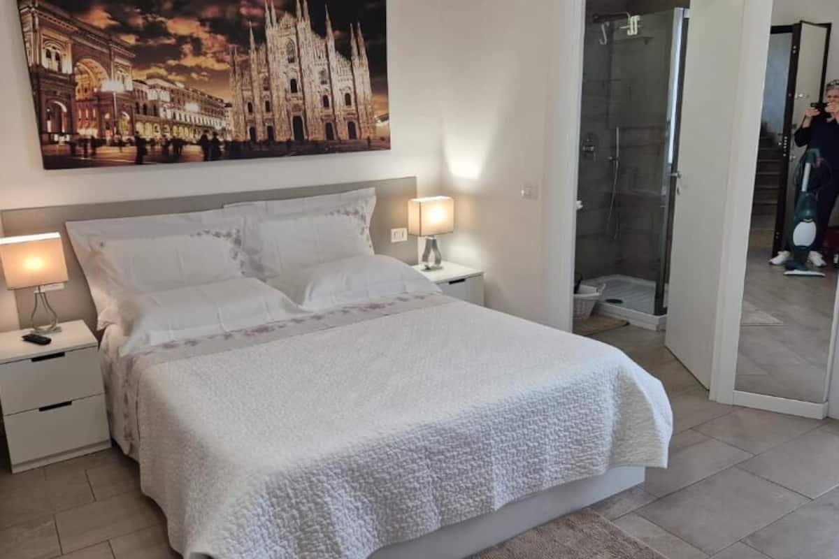 A neatly made bed with white linens, a nightstand with a lamp, and a glass shower enclosure.
