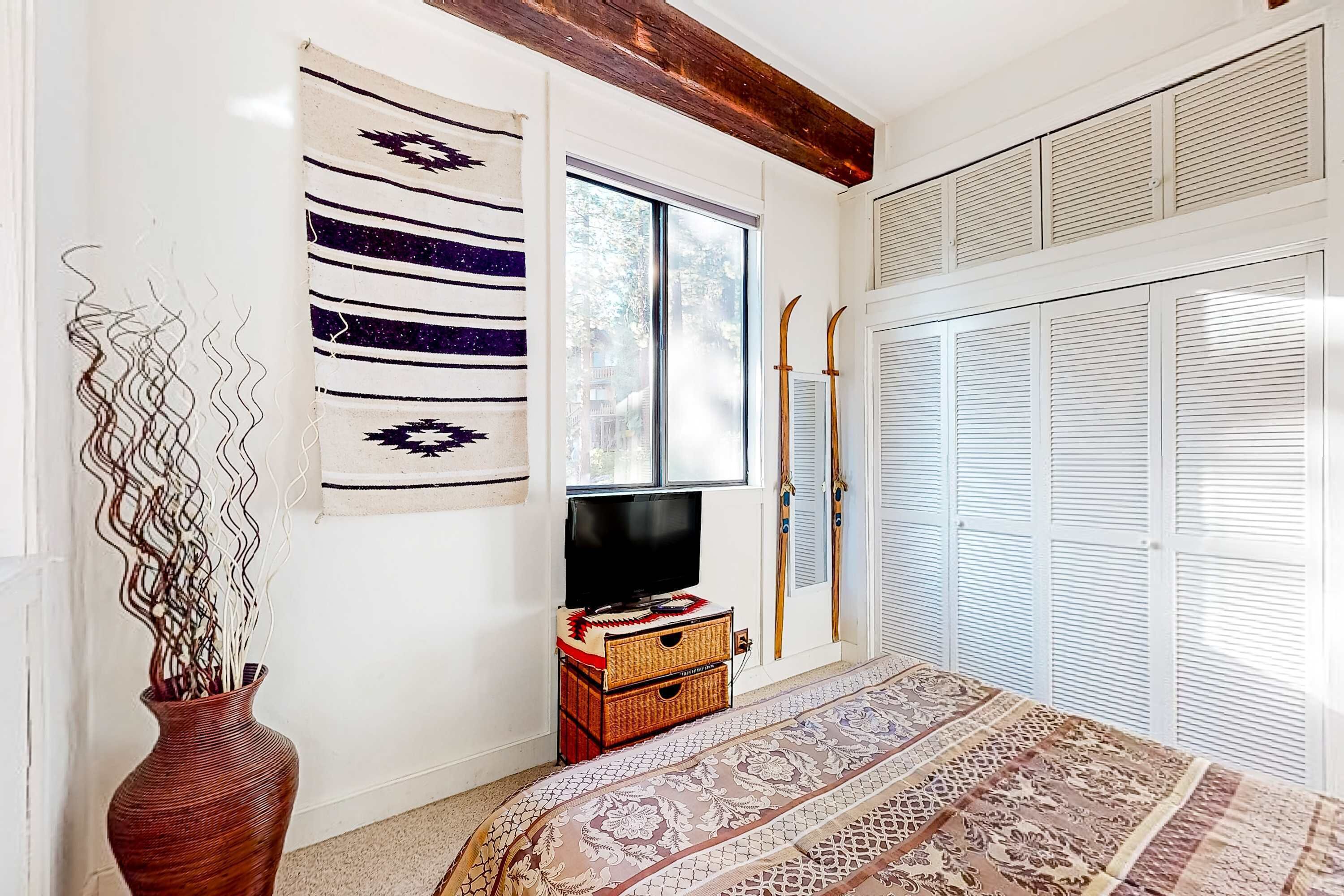 Ein Zimmer mit Fernseher, einem gemusterten Teppich, einem Holzbalken, einem Fenster und einer weißen Tür mit Fensterläden.