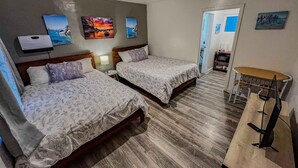 Ein Zimmer mit zwei Betten, einem Schreibtisch und einem Stuhl. An den Wänden befinden sich Kunstwerke, und eine Tür führt in einen weiteren Raum.