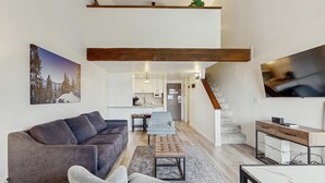 Ein modernes Wohnzimmer mit Sofa, Sessel, Hocker und einem Essbereich mit Tisch und Stühlen. Im Hintergrund ist eine Küche zu sehen, und eine Treppe führt zu einer weiteren Ebene.