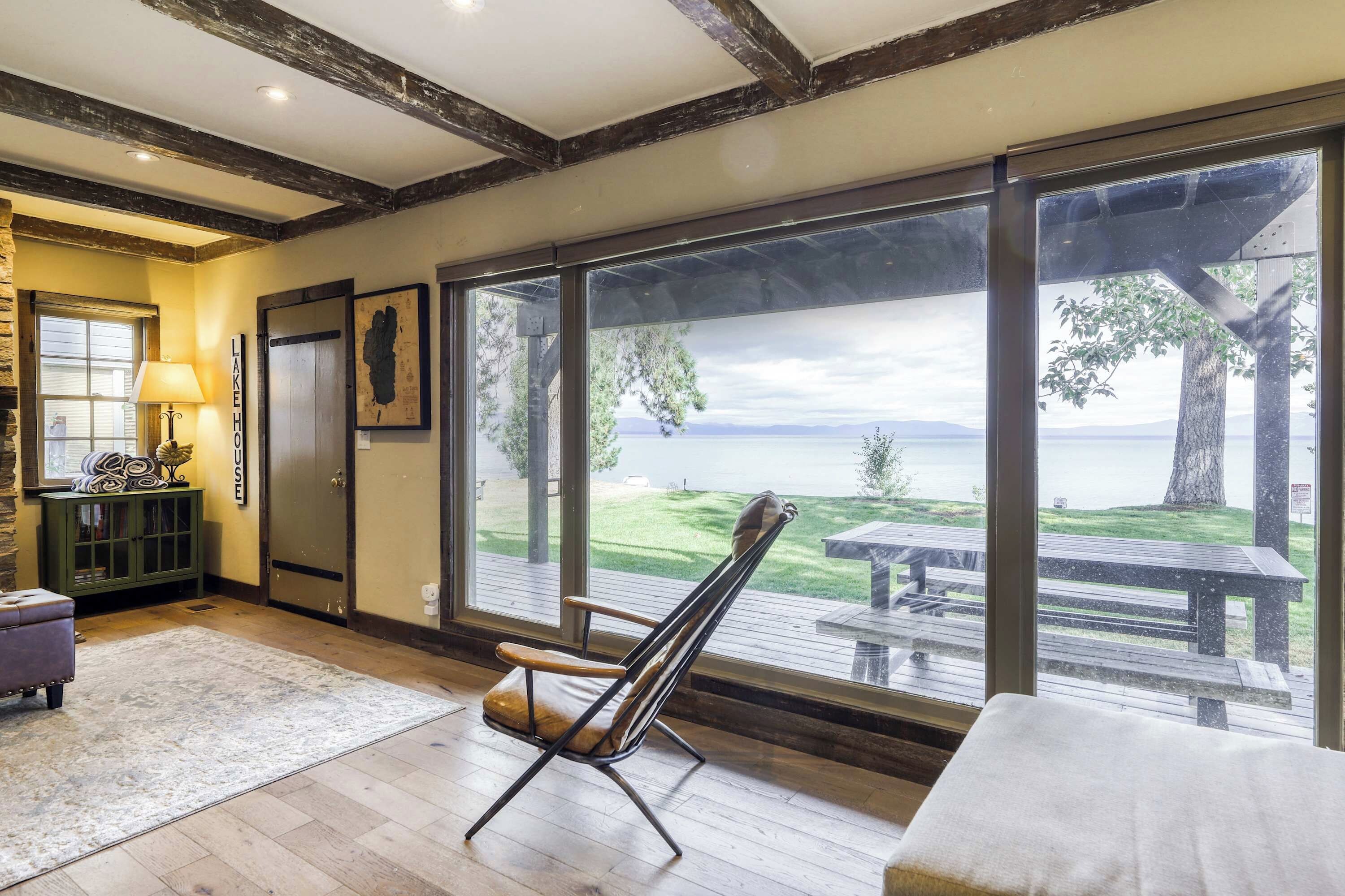 Een kamer met een groot raam dat uitkijkt op een terras en een meer, een stoel, een lamp en een kast.
