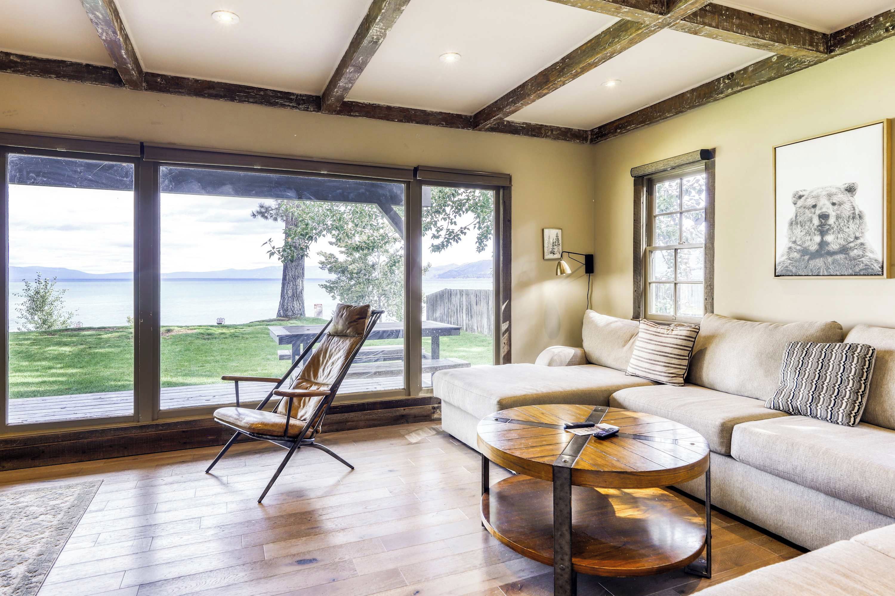 A spacious living room with a large window overlooking a scenic view, a leather chair, a round wooden coffee table, and a sofa with cushions.