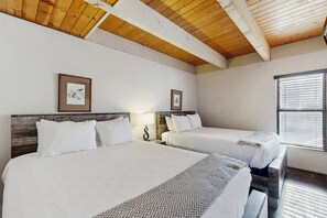 Camera d'albergo con due letti, travi in legno a soffitto, opere d'arte incorniciate alle pareti e una finestra con persiane.