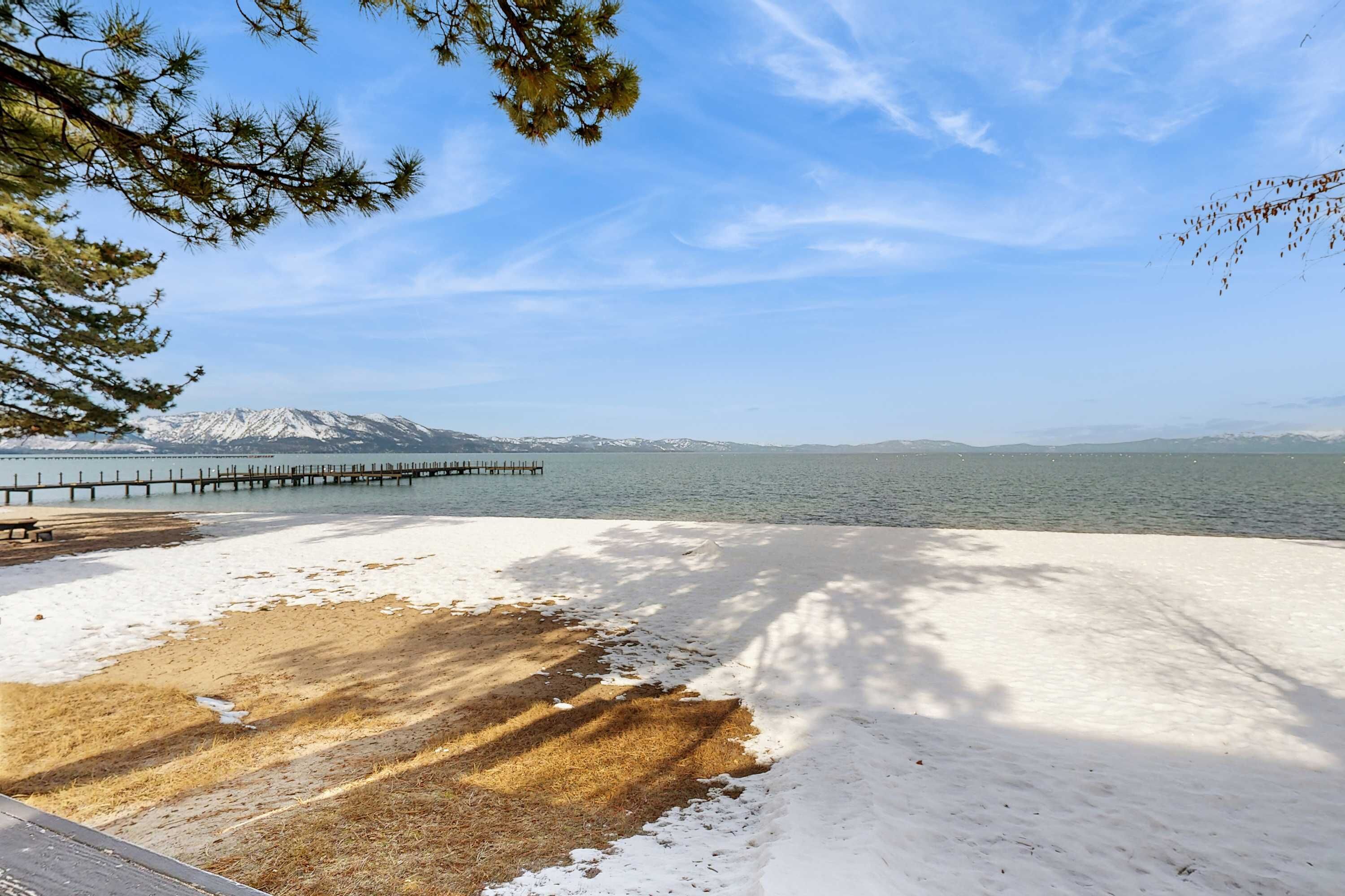 En strand med snöfält, en träbrygga och berg i fjärran.