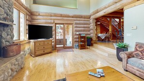 Ein rustikales Blockhaus-Interieur mit Kamin aus Stein, Holzmöbeln, einem Fernseher und einer Treppe, die zu einer erhöhten Ebene führt.