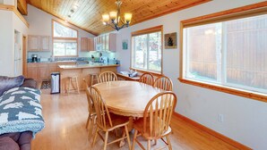 Un interior acogedor con una mesa de comedor de madera, sillas, un sofá y una zona de cocina con armarios de madera y un ventanal grande.
