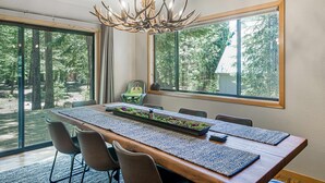 Een eetruimte met een lange houten tafel, stoelen, een kroonluchter en grote ramen met uitzicht op bomen.