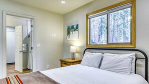 Un dormitorio con una cama, una mesita de noche, una lámpara, una ventana con vistas a árboles y una puerta que da a otra habitación.