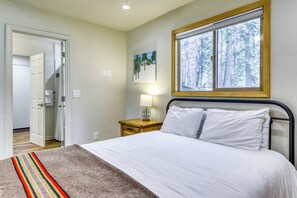 A bedroom with a bed, a nightstand, a lamp, a window with a view of trees, and a door leading to another room. - Charming cabin near downtown Truckee & skiing with large deck (Truckee)