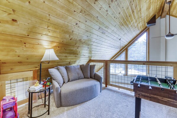Una habitación acogedora con techo de madera, un sofá con cojines, una mesa con una lámpara, una ventana y una mesa de fútbol.