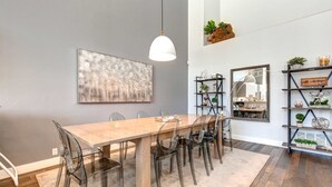 Een eetgedeelte met een houten tafel, transparante stoelen, een hangende hanglamp en een groot abstract schilderij aan de muur.