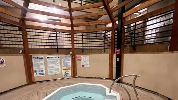 Un spa interior con una ba ñera de hidromasaje hexagonal, vigas de madera y varios letreros de precaución en las paredes.