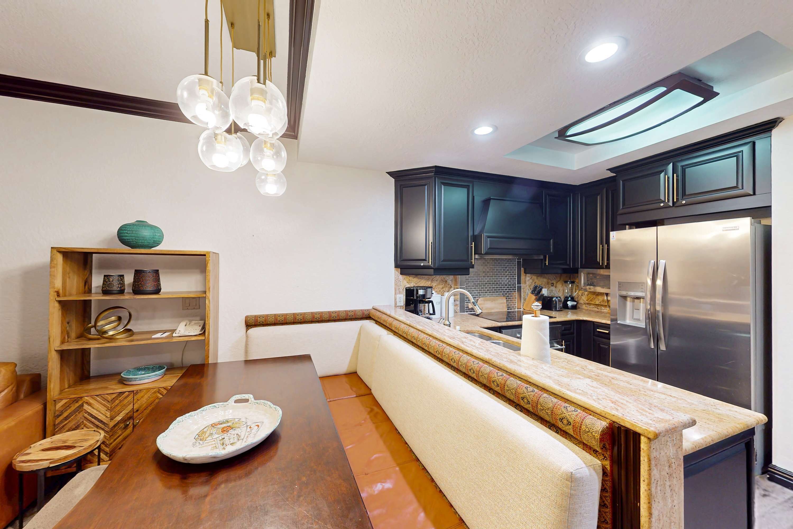 A modern kitchen with dark cabinets, a stainless steel refrigerator, a wooden dining table with a bench, and a hanging light fixture.