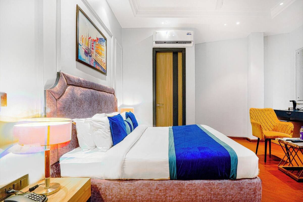 A hotel room with a bed, a desk, a chair, an air conditioning unit, a painting, and a door.