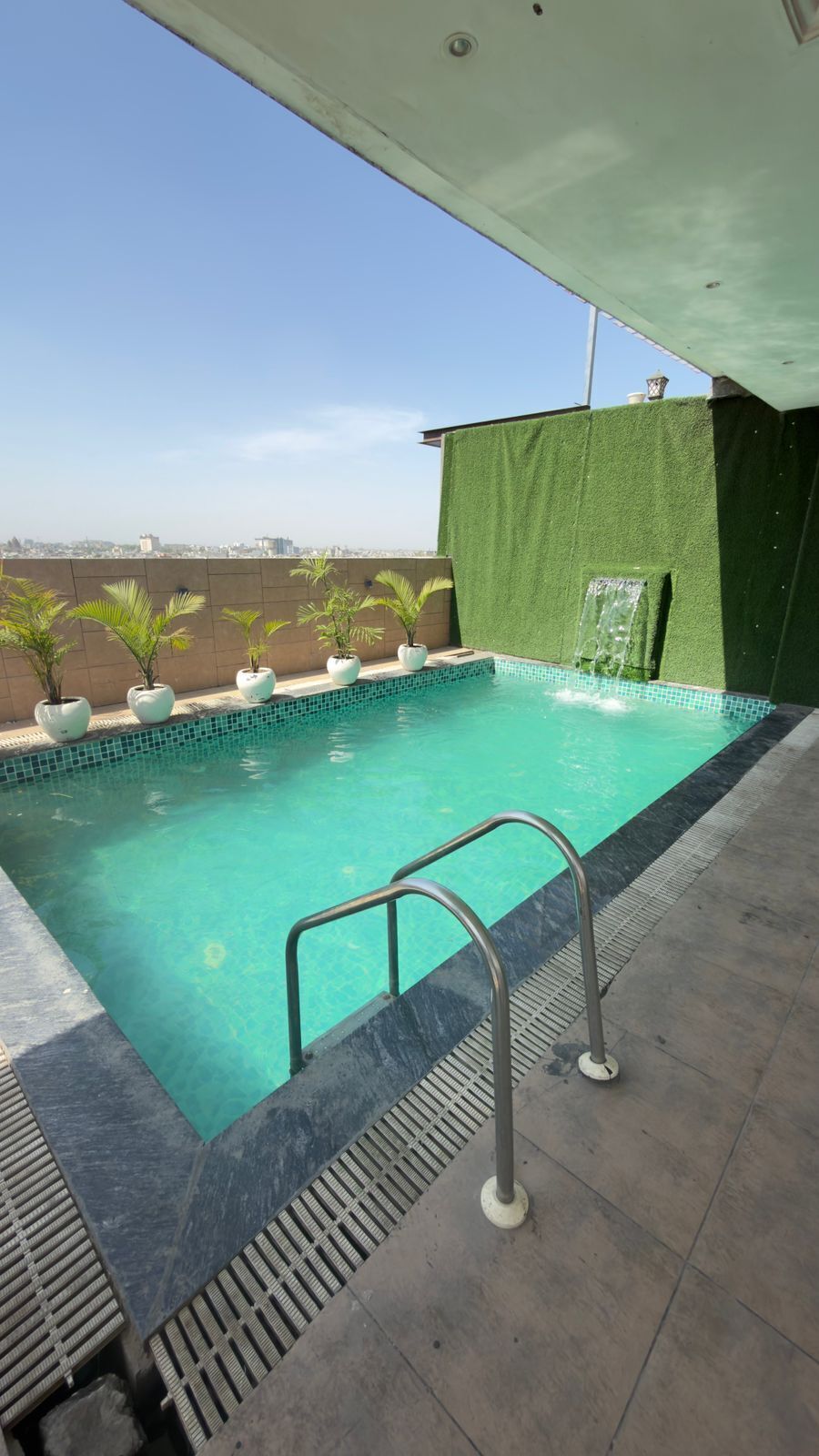 A rooftop pool with a clear view of the cityscape, a green wall, and a small waterfall feature.