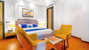 A hotel room with a bed, two chairs, a table, a lamp, a painting, and an air conditioning unit. - Grand Cabbana by White Panda Hotels (Amritsar)