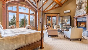 A spacious bedroom with a large bed, a fireplace, and a view of trees through large windows. - Luxury lodge with hot tub, sauna, game room, movie room & Deer Valley views (Park City)