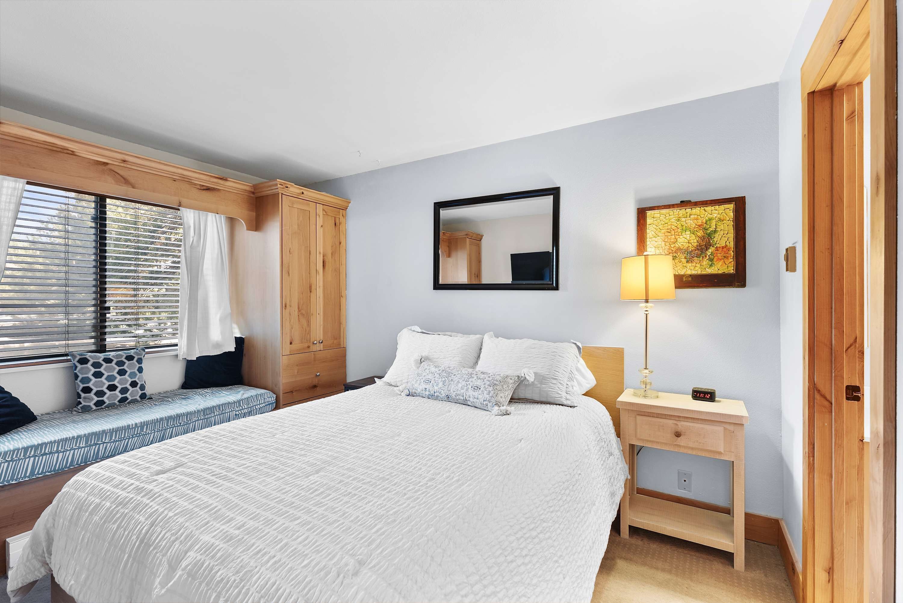Un dormitorio con cama, mesita de noche con lámpara, ventana con persianas, armario de madera y espejo adosado a la pared.