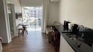 A modern kitchen with a black countertop and stainless steel sink, a dining area with a table and chairs, and a view of buildings through a large window. - Vila do Centro (Bombinhas)