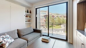 A modern living room with a grey sofa, a coffee table, and a large window with a view of buildings and greenery. - Walk-to-lift studio with hot tub, sauna, pool, gym, & game room - Dog friendly (Park City)