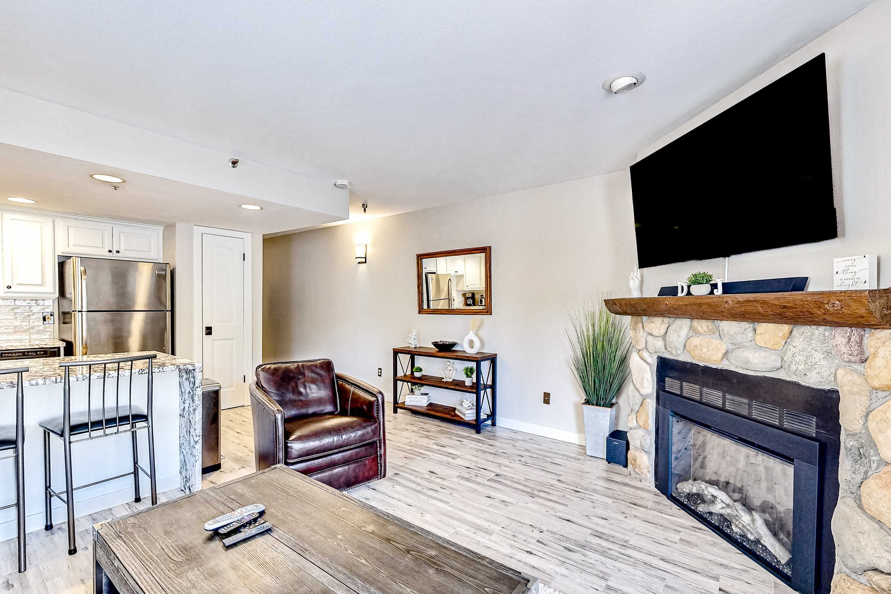 A modern living room with a fireplace, a TV mounted on the wall, a kitchen area with a refrigerator, and a seating area with a leather chair and a coffee table.