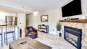 A modern living room with a fireplace, a TV mounted on the wall, a kitchen area with a refrigerator, and a seating area with a leather chair and a coffee table. - Updated condo w/ ski & mountain views, walk to slopes, free Wifi (Park City)