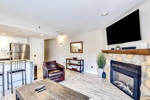 Ein modernes Wohnzimmer mit Kamin, einem an der Wand befestigten Fernseher, einer Küchenzeile mit Kühlschrank sowie einer Sitzecke mit Ledersessel und Couchtisch.