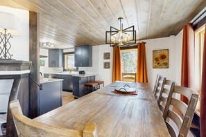 A dining area with a wooden table and chairs, a kitchen in the background, and a ceiling with exposed beams. - Newly remodeled ski-in - ski-out cabin with washer & dryer, new kitchen - walk to lifts (Brighton)